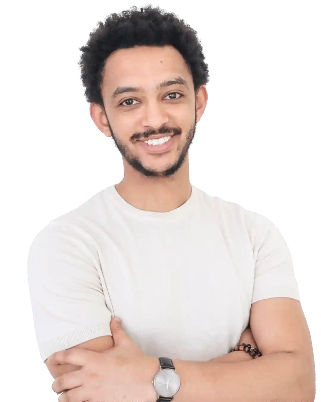 A young man with short curly hair and a beard smiles with arms crossed. He is wearing a light-colored t-shirt, a wristwatch, and a beaded bracelet, standing against a plain background.