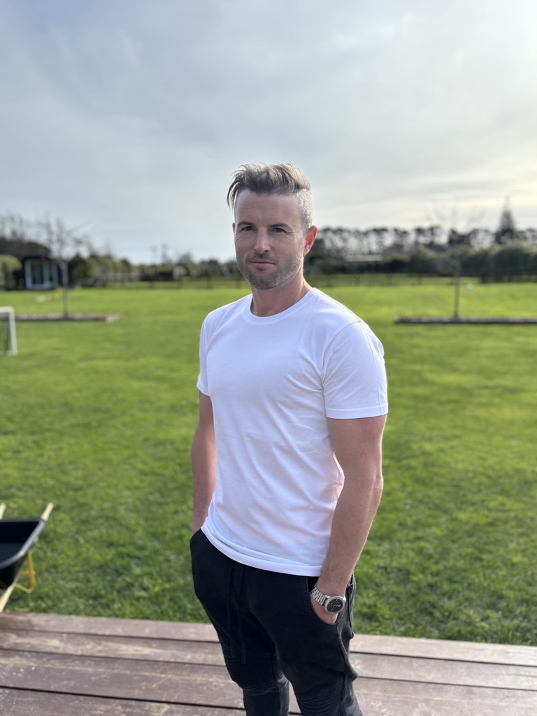 A man with short, styled hair wearing a white t-shirt and black pants stands outdoors on wooden decking, with green grass and trees in the background under a bright, slightly cloudy sky.