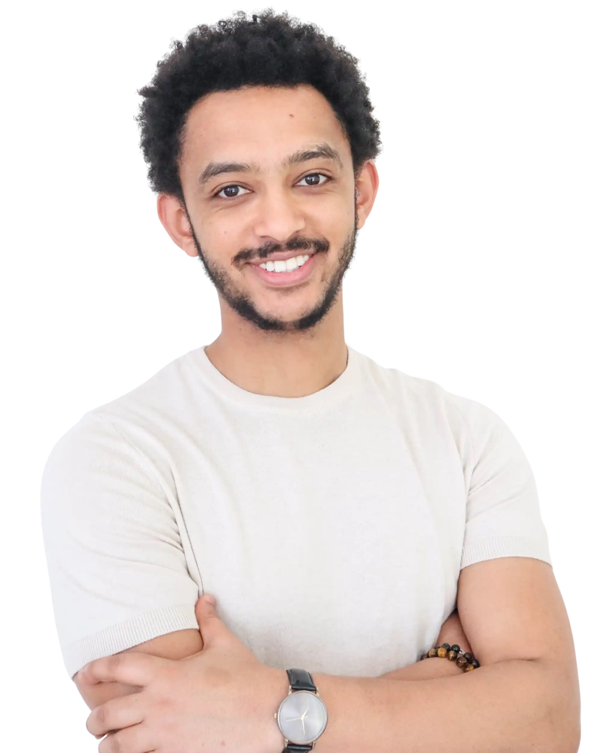 A smiling man with short curly hair and a beard stands against a white background, arms crossed. He wears a light-colored short-sleeve shirt, a wristwatch, and a beaded bracelet.