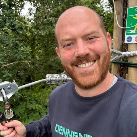 A smiling person with a trimmed beard stands outdoors near zipline equipment, wearing a dark sweatshirt. Green trees and a safety sign are visible in the background.